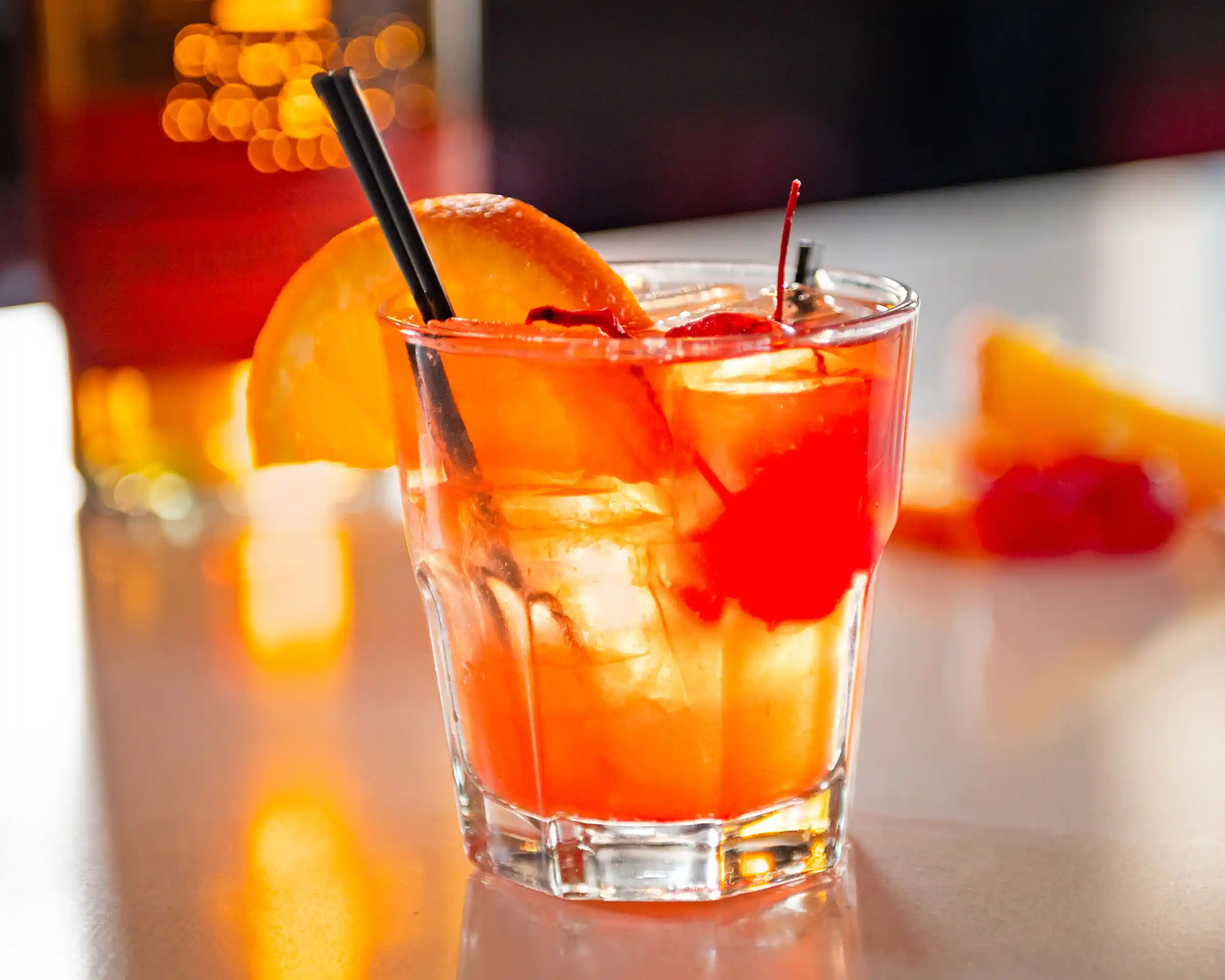 Orange-red cocktail with ice, orange slice, and cherry garnish.